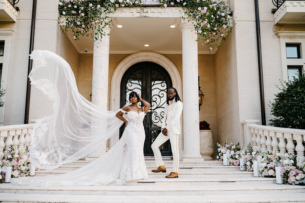 Bride’s cathedral veil billows dramatically beside the couple standing on the front steps of a floral-adorned mansion entrance.