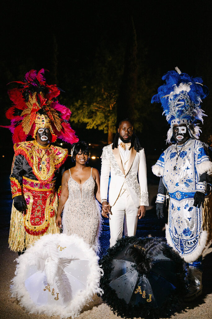 Bride and groom stand between two elaborately dressed Mardi Gras Indians during a nighttime wedding portrait in New Orleans.