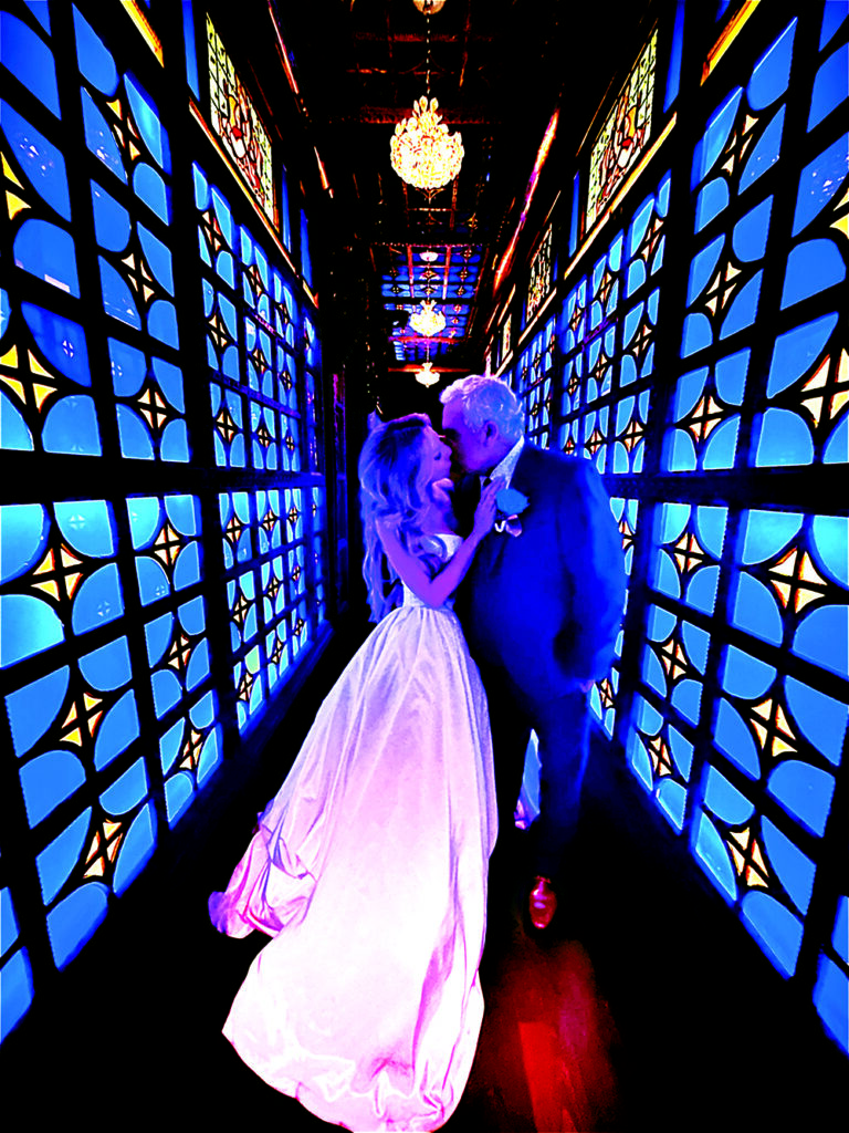 Bride and groom kiss beneath glowing chandeliers in a dramatic blue-lit corridor lined with stained-glass-inspired panels at their masquerade-themed wedding celebration.