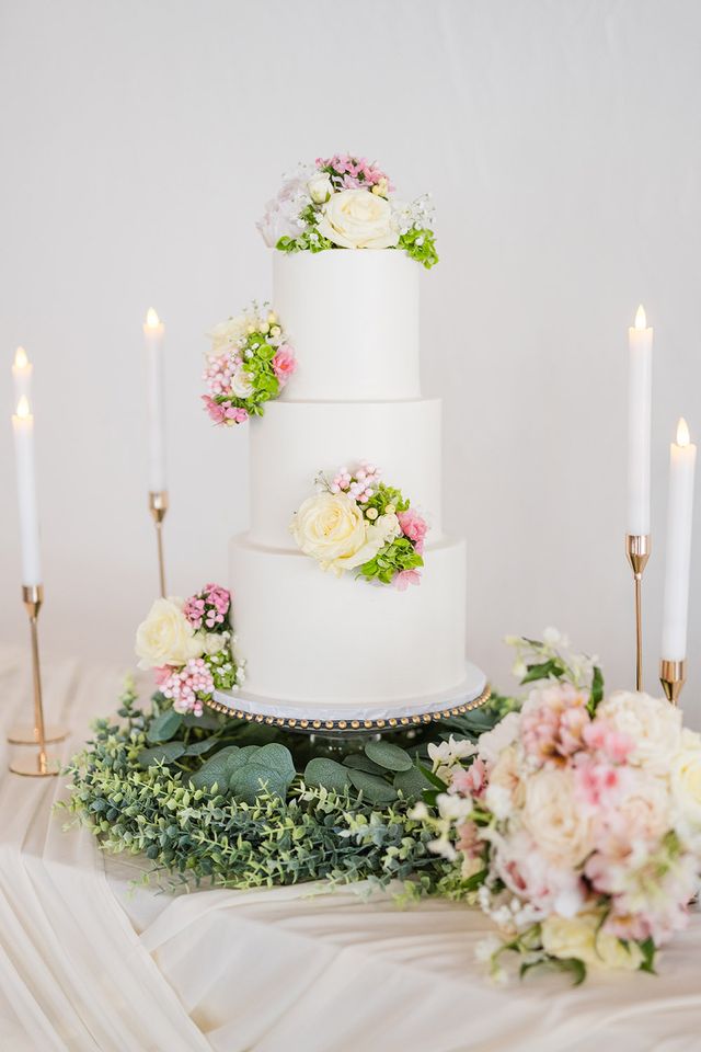 Three-tier white wedding cake with pastel floral accents and gold candle holders on elegant display table