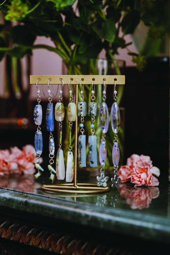 Handmade gemstone earrings displayed on a gold stand beside soft pink flowers and blurred greenery in the background.