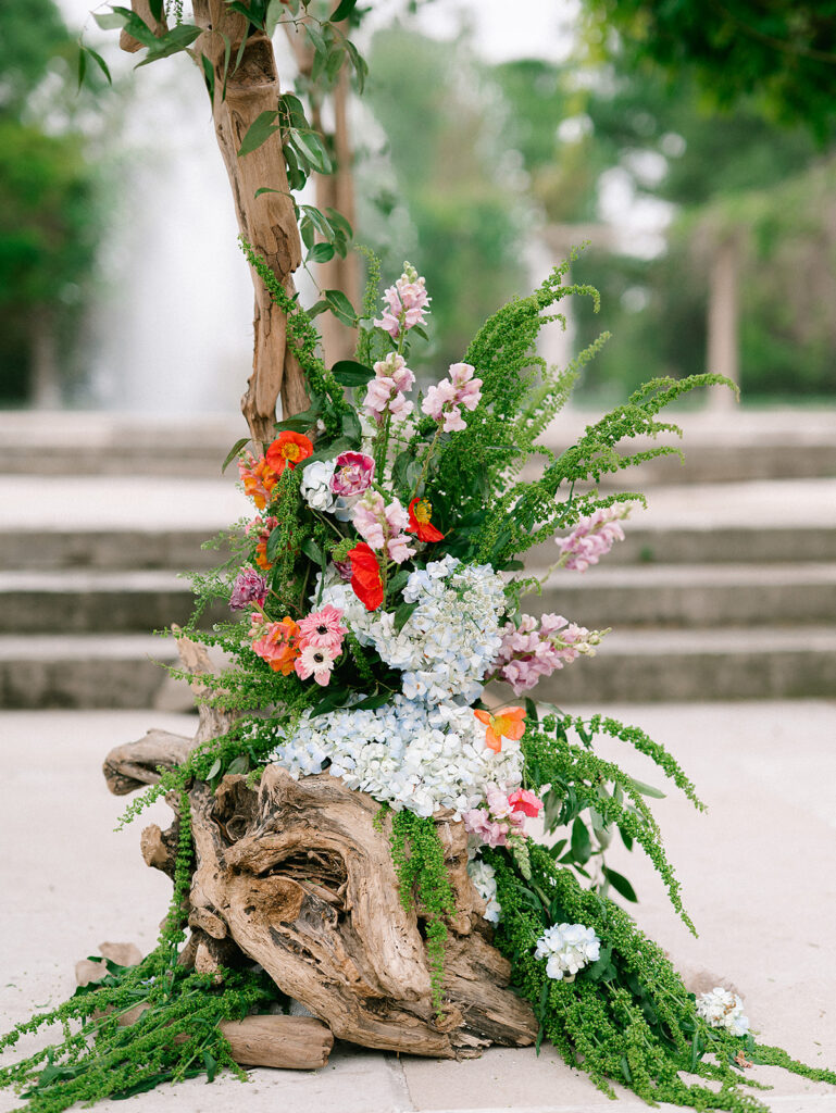 Whimsical floral installation of hydrangeas, snapdragons, greenery, and bright blooms arranged around driftwood near a fountain.
