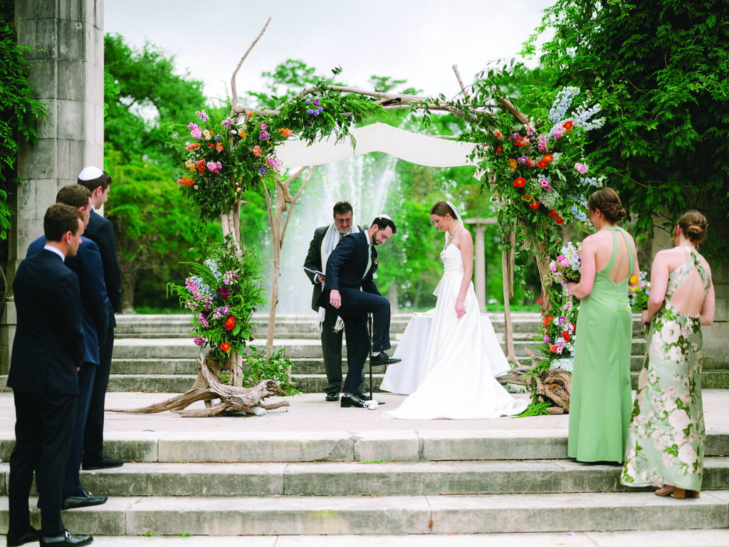 Bride and groom stand beneath a lush floral chuppah during an outdoor wedding ceremony surrounded by family and wedding party members.