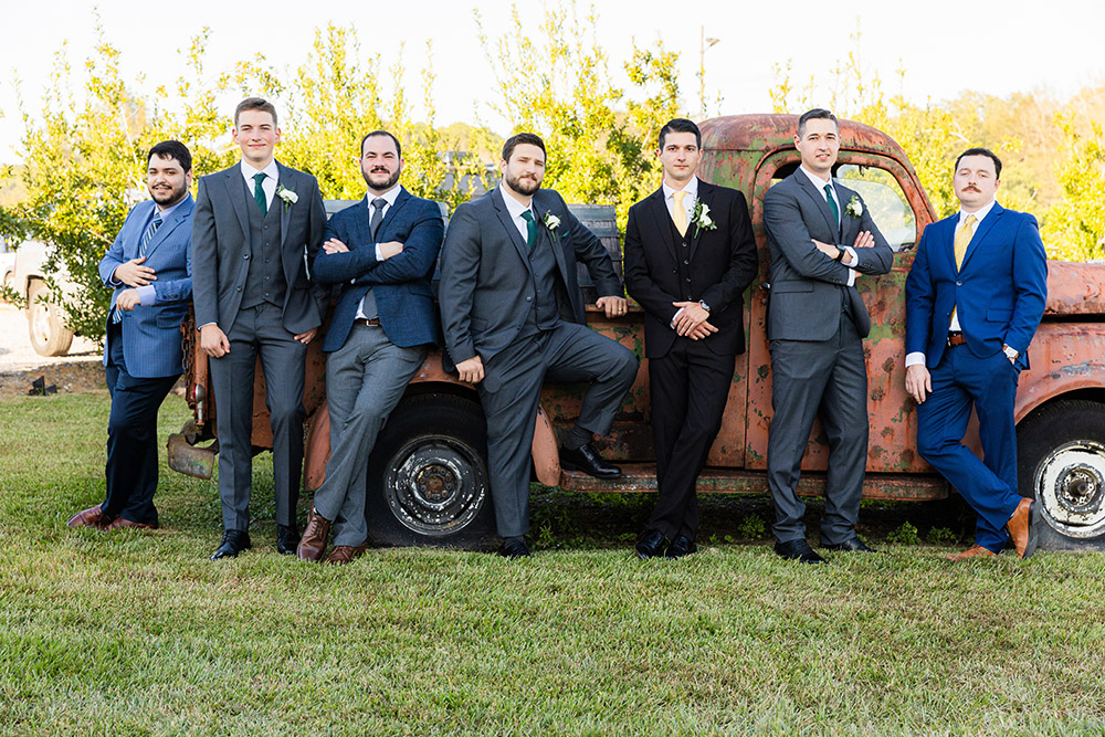 Groom and groomsmen posing in suits beside a vintage rust-colored pickup truck outdoors before the ceremony.