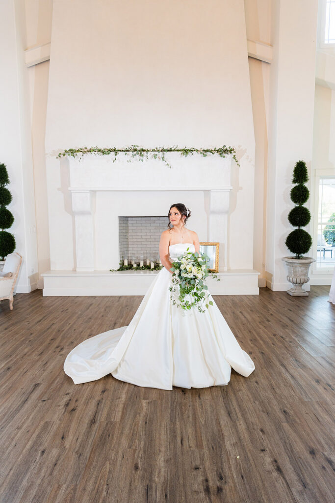 Bride in a strapless satin ball gown posing with a cascading white and greenery bouquet inside a bright white reception hall with a grand fireplace backdrop.