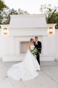 Newlyweds posing in front of a white outdoor fireplace feature, the bride holding a lush greenery bouquet with her satin train flowing across the ground.
