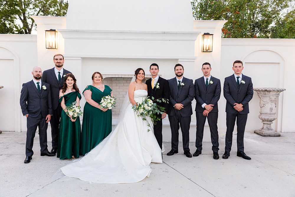 Wedding party portrait with the bride and groom surrounded by bridesmaids in emerald green dresses and groomsmen in dark suits outside the ceremony space.