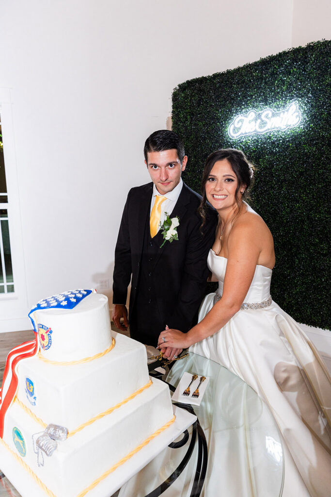 Newlyweds cutting a white tiered wedding cake decorated with military-inspired emblems beside a greenery wall and neon sign at the reception.