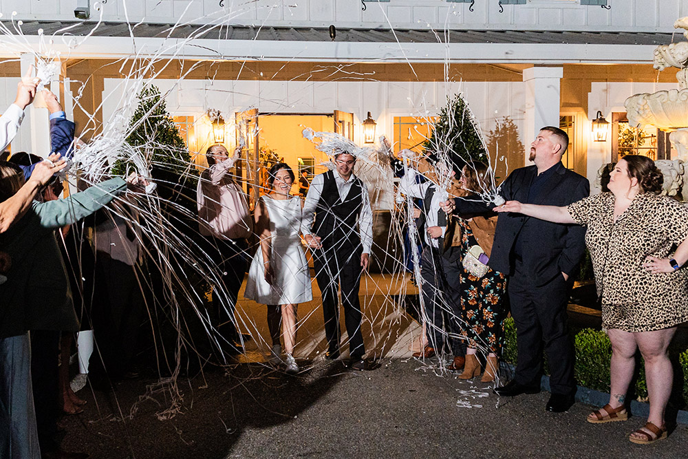 Bride and groom making a festive streamer exit at the end of the reception while guests celebrate around them outdoors at night.