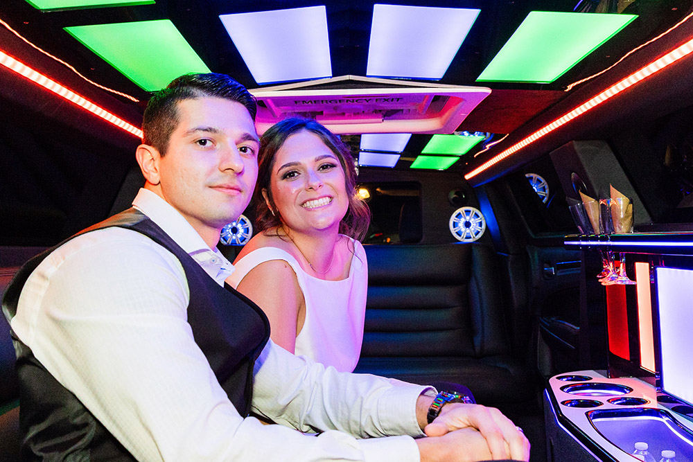 Newlyweds sitting together inside a limousine with colorful neon lighting and champagne glasses after their wedding reception.