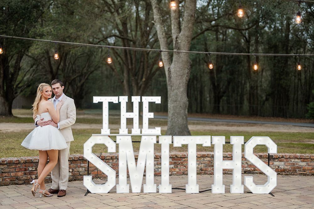 Bride and groom embracing beside large marquee “THE SMITHS” sign with string lights overhead