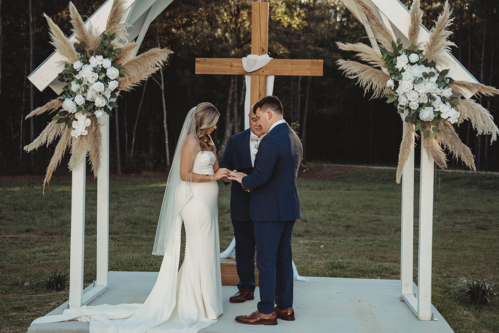 Bride and groom exchange vows beneath a white chapel-style arbor with pampas grass floral arrangements and a large wooden cross.