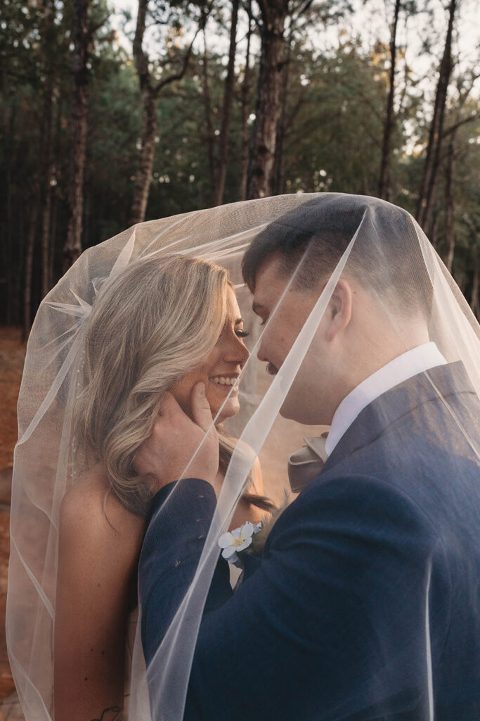 Bride and groom share an intimate moment beneath the veil in a pine forest as the groom gently touches the brideโs face.