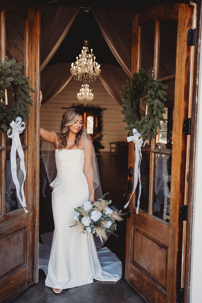 Bride in a fitted strapless wedding gown stands in the doorway of the chapel holding a bouquet of white roses and pampas grass beneath crystal chandeliers.