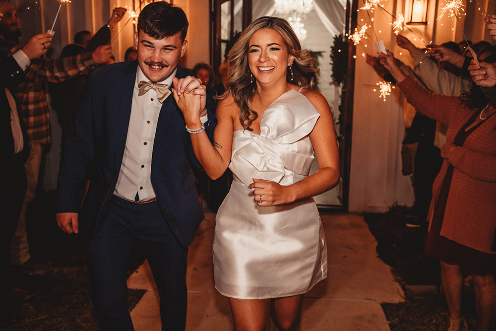 Newlyweds Seth and Ashley smile while walking through a sparkler exit, hand in hand, as guests celebrate around them at their evening wedding reception.