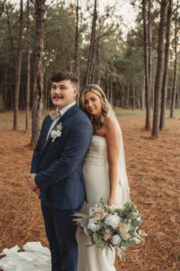 Bride and groom pose together in a pine forest, with the bride holding a bouquet of white roses, eucalyptus, and pampas grass.