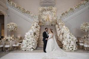 Bride and groom stand beneath grand floral staircase installation with cascading white flowers and monogrammed wedding cake display in elegant reception ballroom