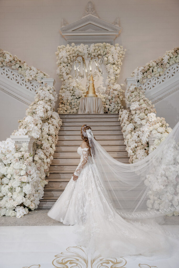 Bride in floral appliqué ball gown with dramatic cathedral veil posing on staircase surrounded by lush white floral arrangements and wedding cake display