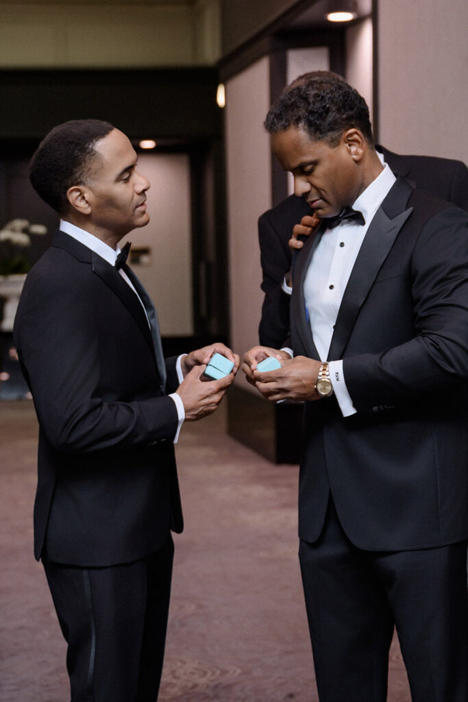 Groom and groomsman in classic black tuxedos opening Tiffany & Co. gift boxes in hotel hallway before wedding ceremony