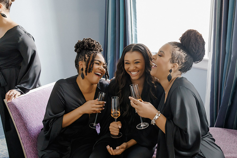 Bridesmaids in black satin robes laughing and toasting champagne while seated together during bridal suite preparations