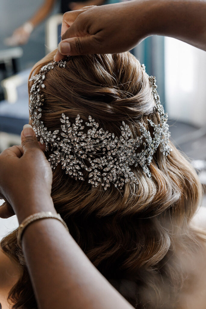 Hairstylist placing sparkling crystal bridal hairpiece into bride’s softly curled half-up wedding hairstyle
