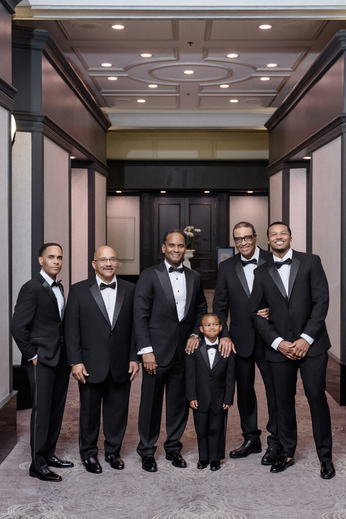 Groom poses with father, grandfather, groomsmen, and young ring bearer in matching black tuxedos in elegant hotel hallway