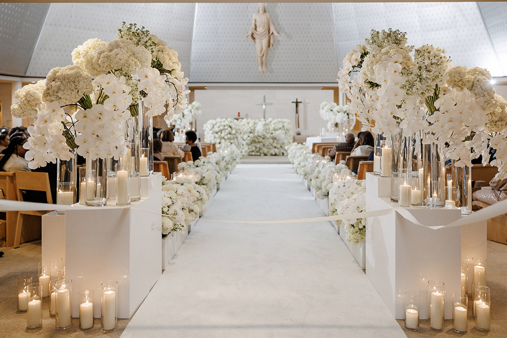 Luxurious white floral wedding aisle lined with candles and orchids inside modern church ceremony space beneath large statue of Christ