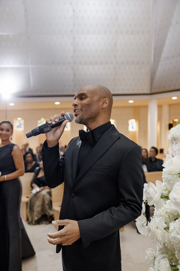 Musician in a black tuxedo sings into a microphone during the wedding ceremony as guests look on from wooden pews.