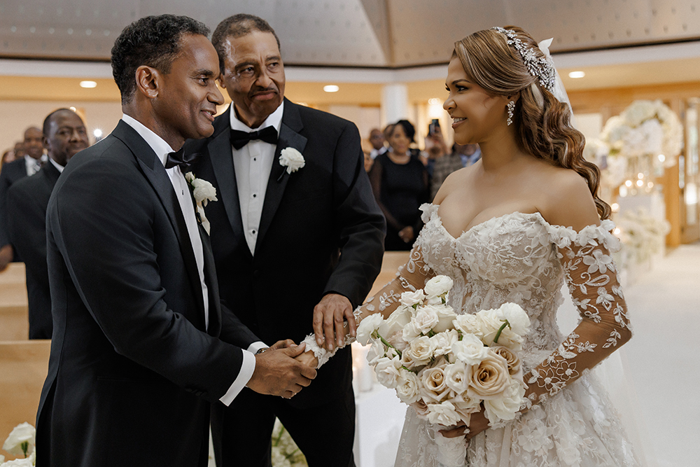 Bride in an off-the-shoulder floral lace wedding gown smiles while joining hands with the groom during the ceremony.