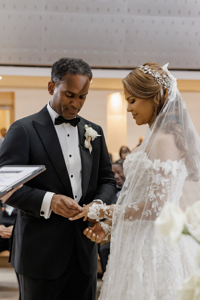 Groom places a wedding band on the bride’s finger during an intimate ceremony surrounded by white floral arrangements.
