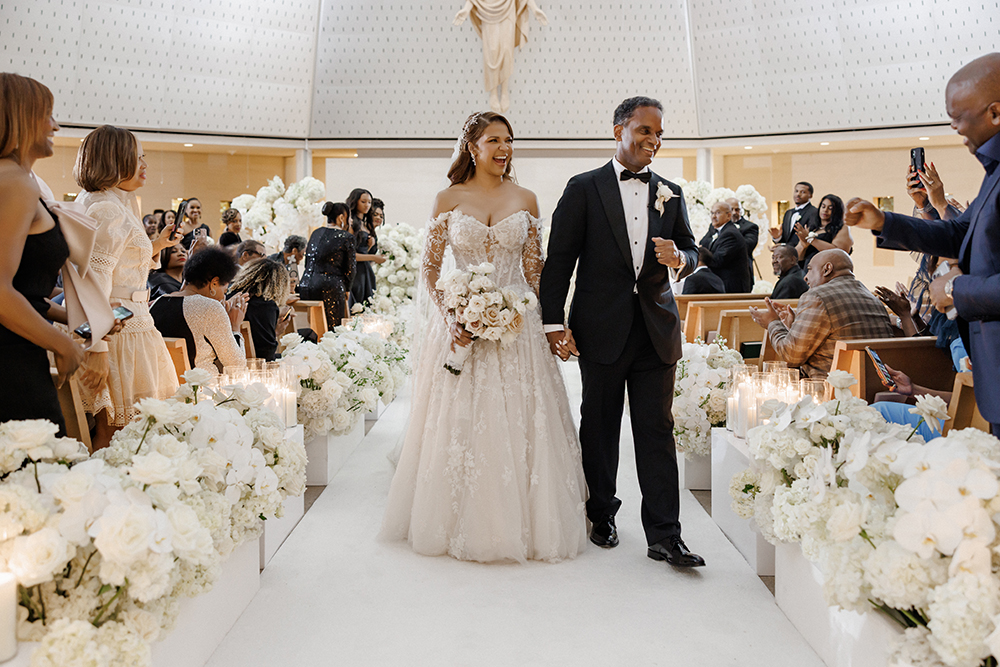 Newlyweds walk down a flower-lined aisle hand in hand after the ceremony as guests applaud from their seats.
