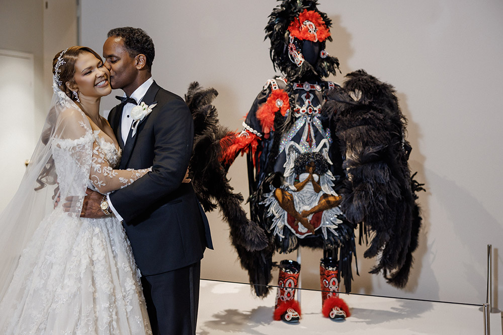 Bride and groom embrace beside a vibrant feathered Mardi Gras Indian-inspired art piece inside the museum venue.