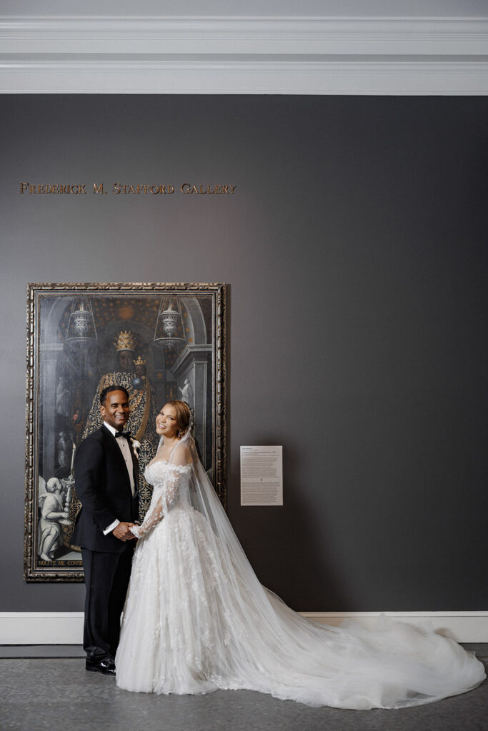 Bride and groom pose beneath the “Frederick M. Stafford Gallery” sign with the bride’s cathedral-length veil trailing behind her.