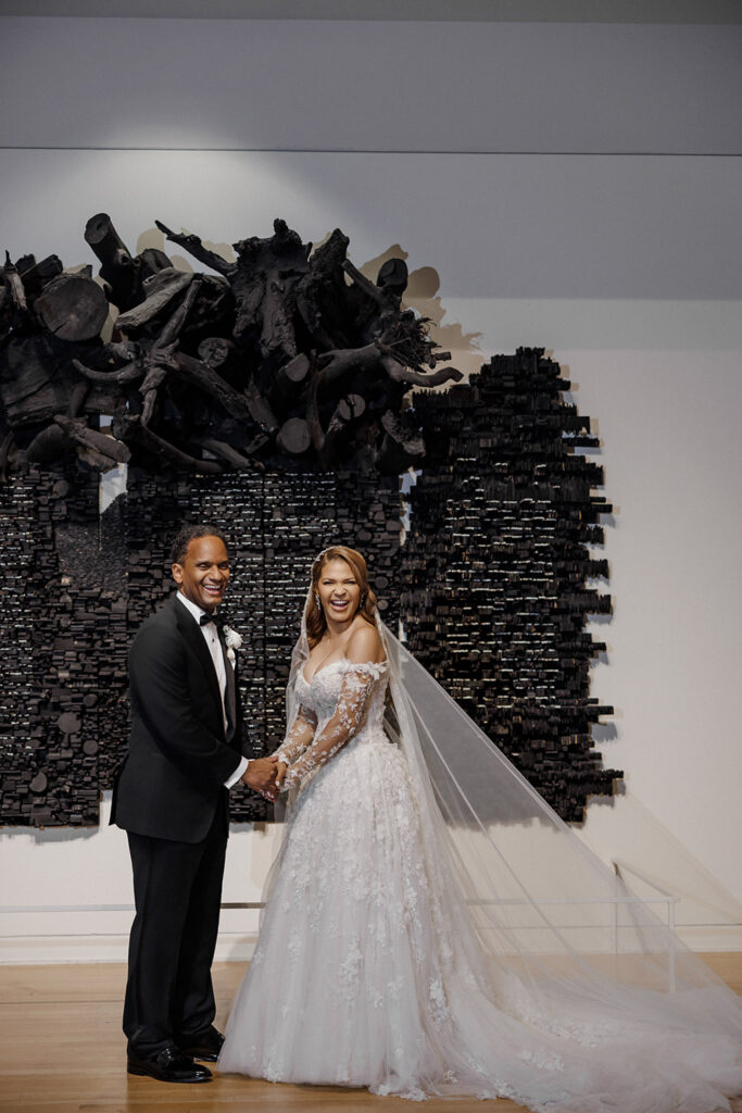Bride and groom laugh together while posing in front of a dramatic black sculptural art installation inside the museum.