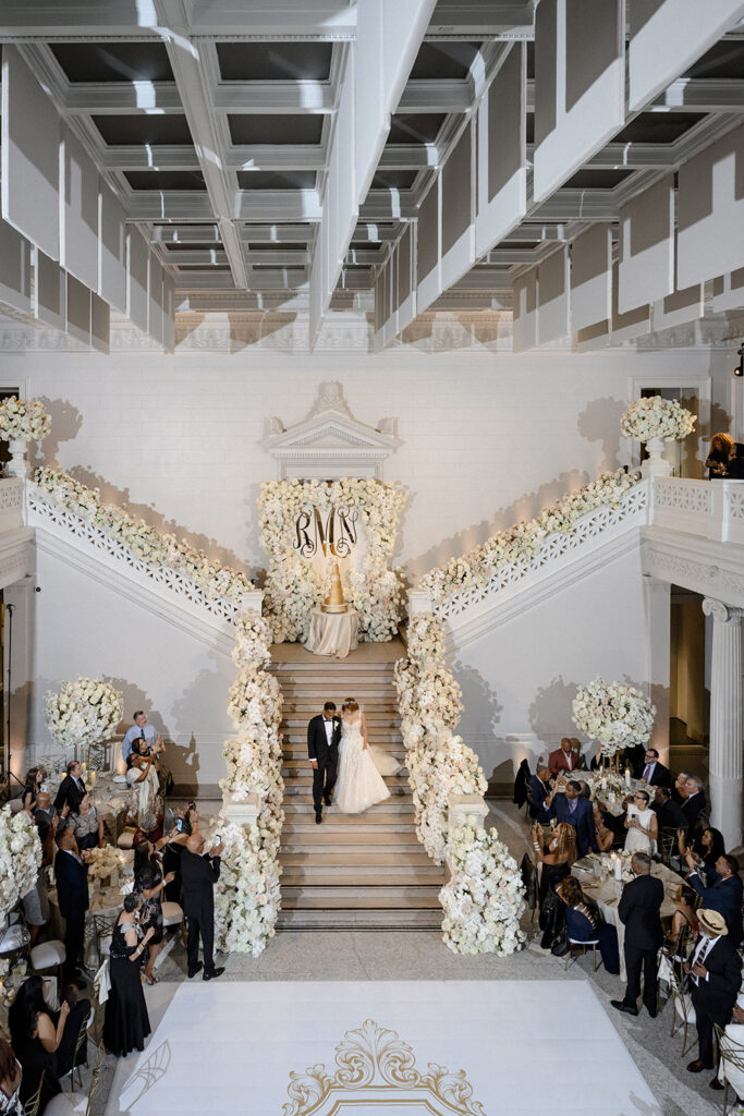 Grand staircase reception entrance covered in lush white flowers with the couple’s monogram displayed at the center landing.