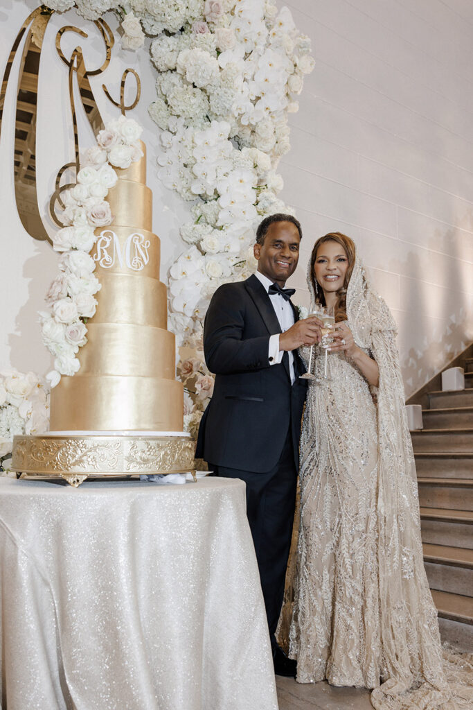 Bride and groom raise champagne glasses beside a towering gold wedding cake framed by lush white floral arrangements and orchids.
