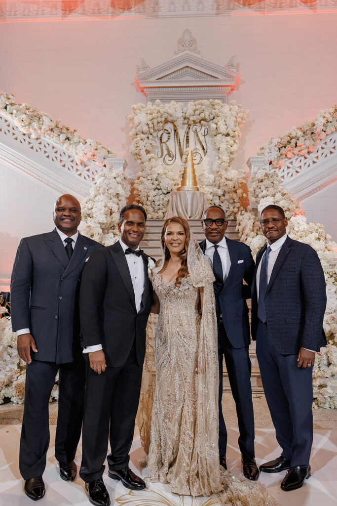 Bride and groom pose with family members in formal attire beneath the grand floral staircase and gold monogram backdrop.