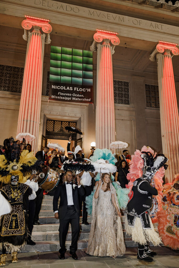 Bride and groom lead a vibrant second line celebration outside the museum surrounded by Mardi Gras Indians, parasols, and brass band musicians.