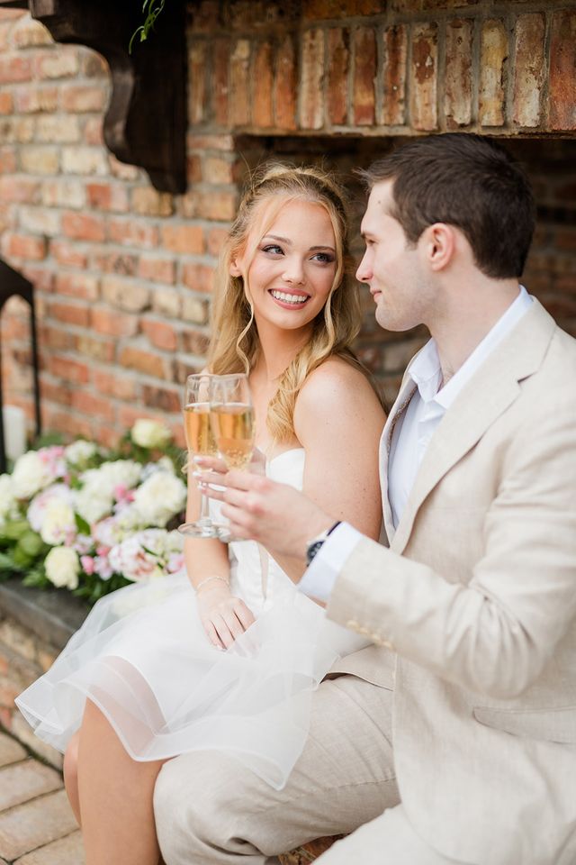 Bride and groom toasting champagne while seated by rustic brick fireplace with floral arrangements