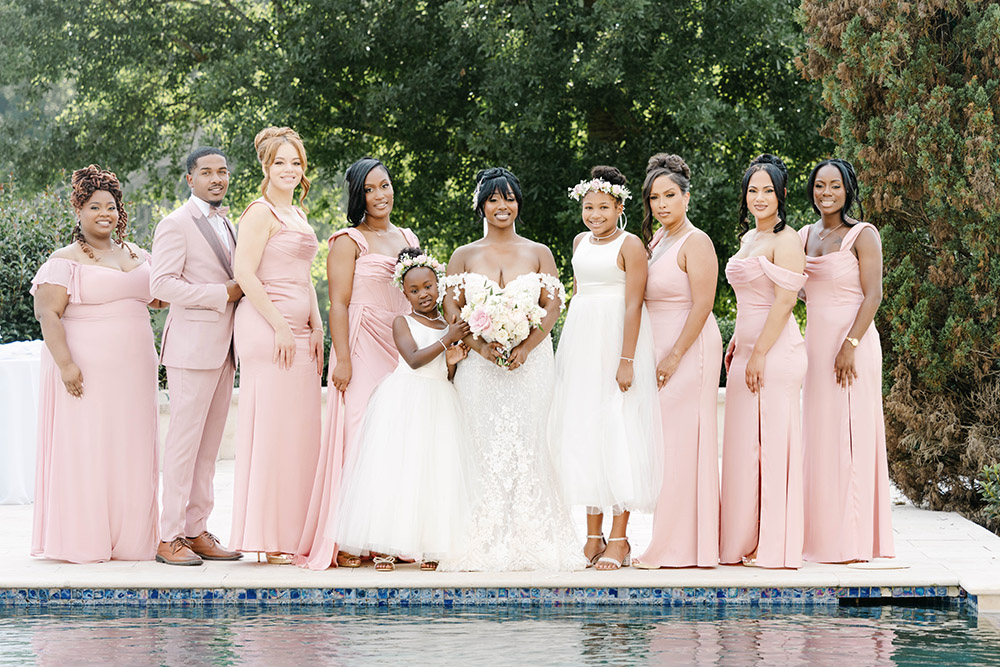 Bride poses with bridesmaids, flower girls, and a groomsman beside the pool in coordinated blush pink dresses and soft romantic wedding attire.