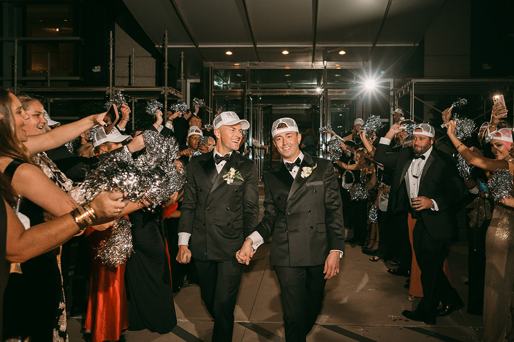Two grooms in black tuxedos walk hand in hand through a festive reception exit as guests cheer with silver pom-poms and matching white party hats.