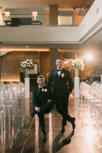 The grooms pose together in the ceremony space surrounded by white floral arrangements, warm lighting, and rows of clear acrylic chairs.