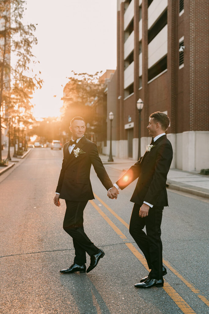 Ryan and Colt hold hands while crossing a downtown Mobile street at sunset, captured in golden light in their classic black tuxedos.
