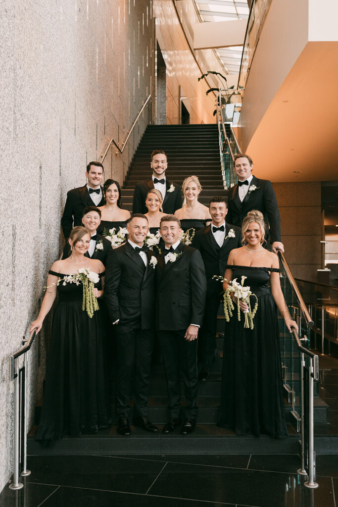 Ryan and Colt pose with their wedding party on a grand staircase, surrounded by bridesmaids in black gowns and white floral bouquets.