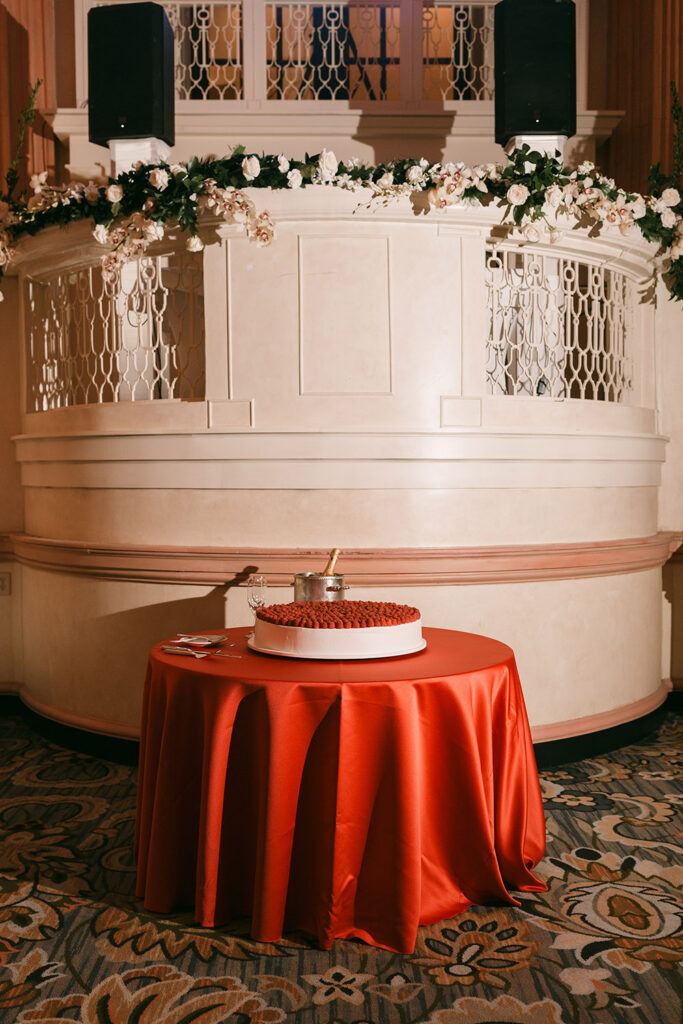A modern white cake topped with fresh raspberries sits on a dramatic red linen table beneath romantic reception lighting and floral garlands.
