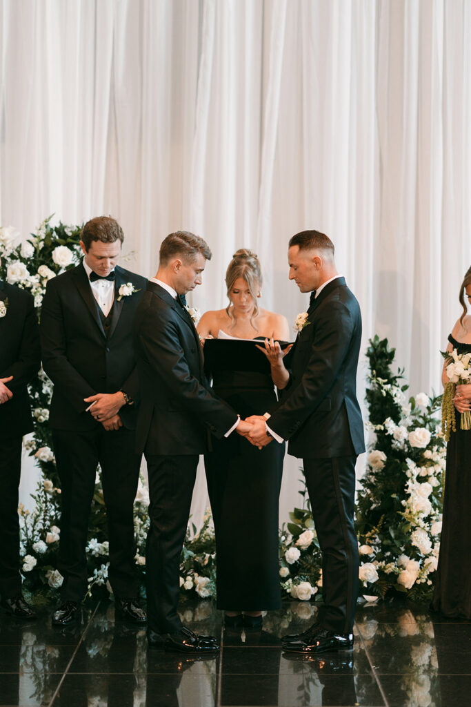 Ryan and Colt hold hands during their wedding ceremony surrounded by lush white floral arrangements and loved ones dressed in black formalwear.