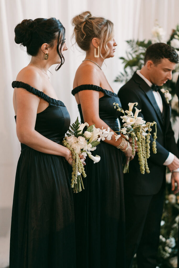 Bridesmaids in black off-the-shoulder gowns hold cascading white bouquets with orchids, roses, and hanging greenery during the ceremony.