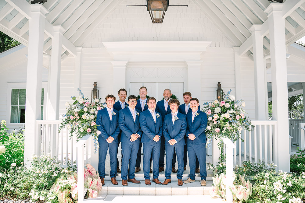 Groom and groomsmen in matching blue suits pose on the steps of a white chapel framed by pastel floral arrangements.
