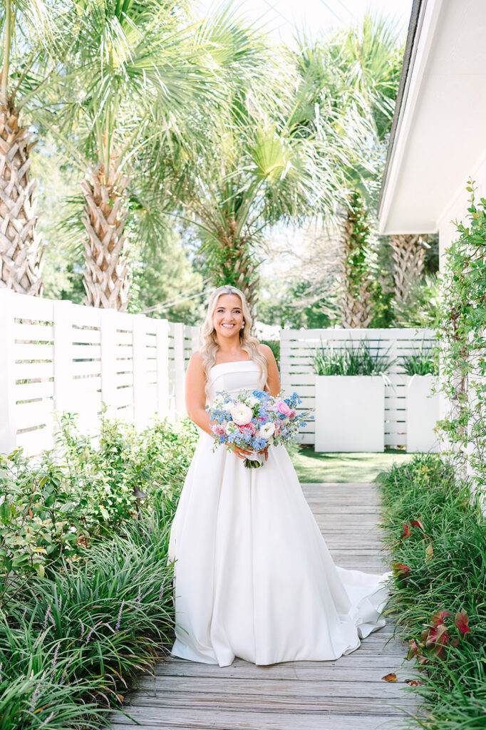 Bride in a strapless satin wedding gown holding a pastel blue and pink bouquet while standing along a palm-lined garden walkway.