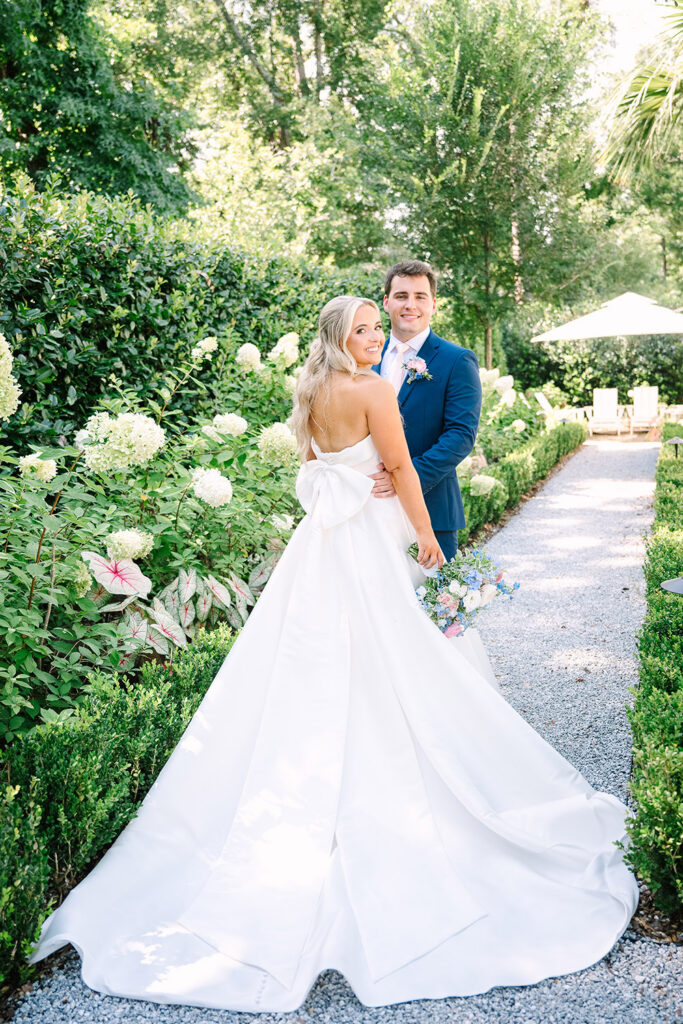 Bride and groom embrace in a garden pathway as the brideโs satin gown with oversized back bow flows behind them.